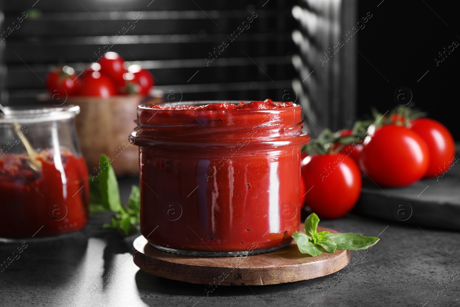 Photo of Jar of tasty tomato paste and basil on grey textured table, closeup