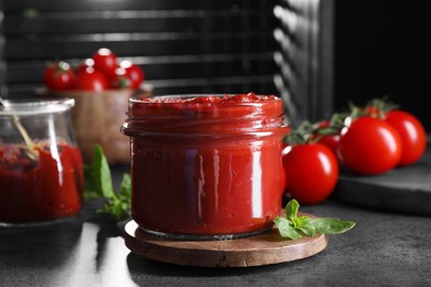 Photo of Jar of tasty tomato paste and basil on grey textured table, closeup