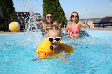 Little girl swimming with inflatable ring near her parents in outdoor pool on sunny summer day