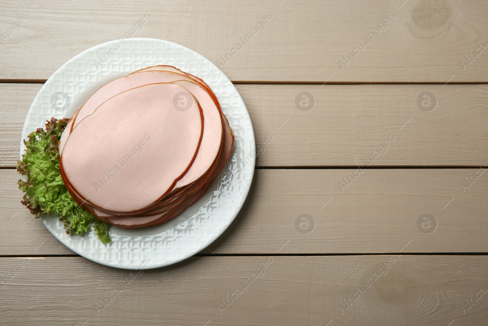 Photo of Slices of delicious boiled sausage with lettuce on beige wooden table, top view. Space for text