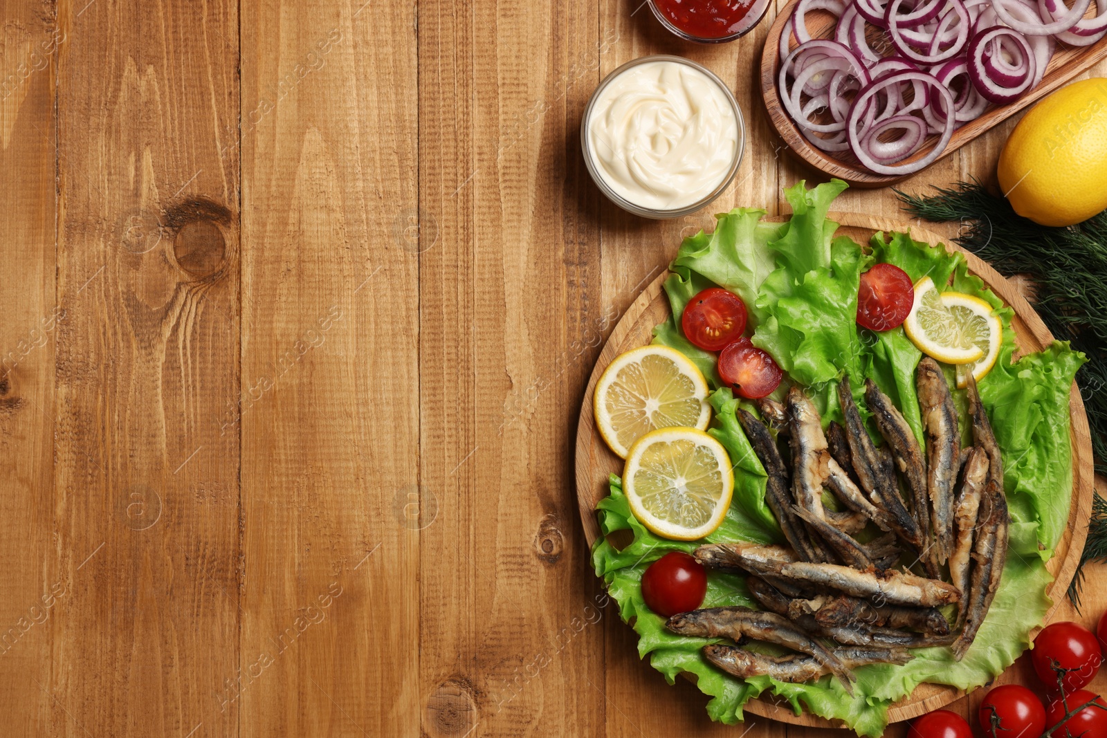 Photo of Delicious fried anchovies served with lemon, tomatoes and sauce on wooden table, flat lay. Space for text