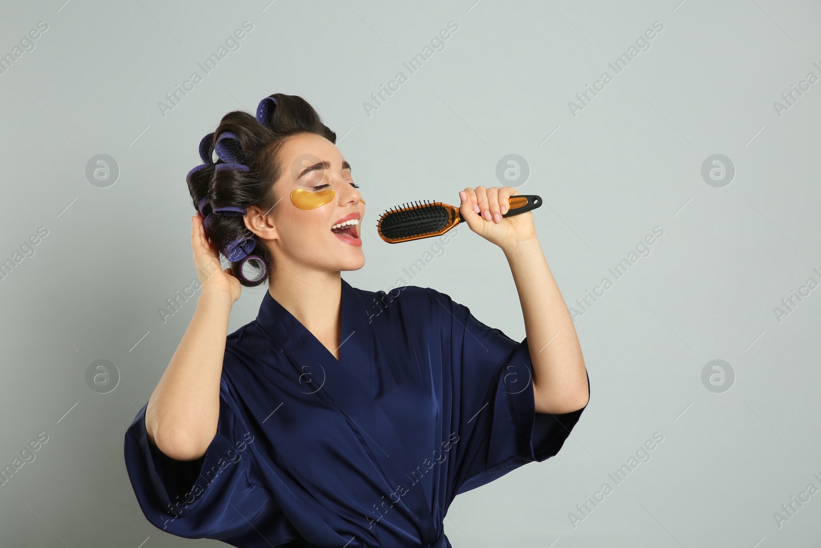 Photo of Happy young woman in silk bathrobe with hair curlers and eye patches singing into hairbrush on grey background, space for text