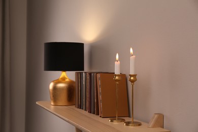 Photo of Wooden shelf with different books, burning candles and lamp on light wall