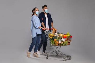Photo of Couple with protective masks and shopping cart full of groceries on light grey background