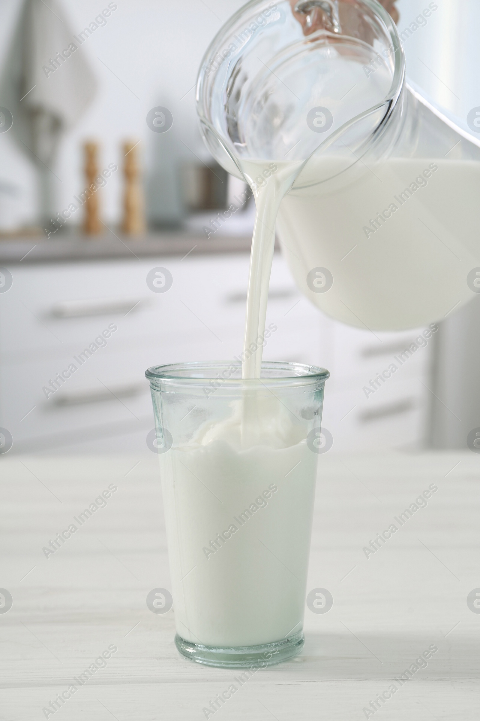 Photo of Woman pouring milk from jug into glass at white wooden table in kitchen, closeup