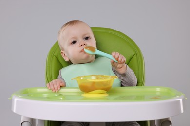 Cute little baby eating healthy food in high chair on gray background