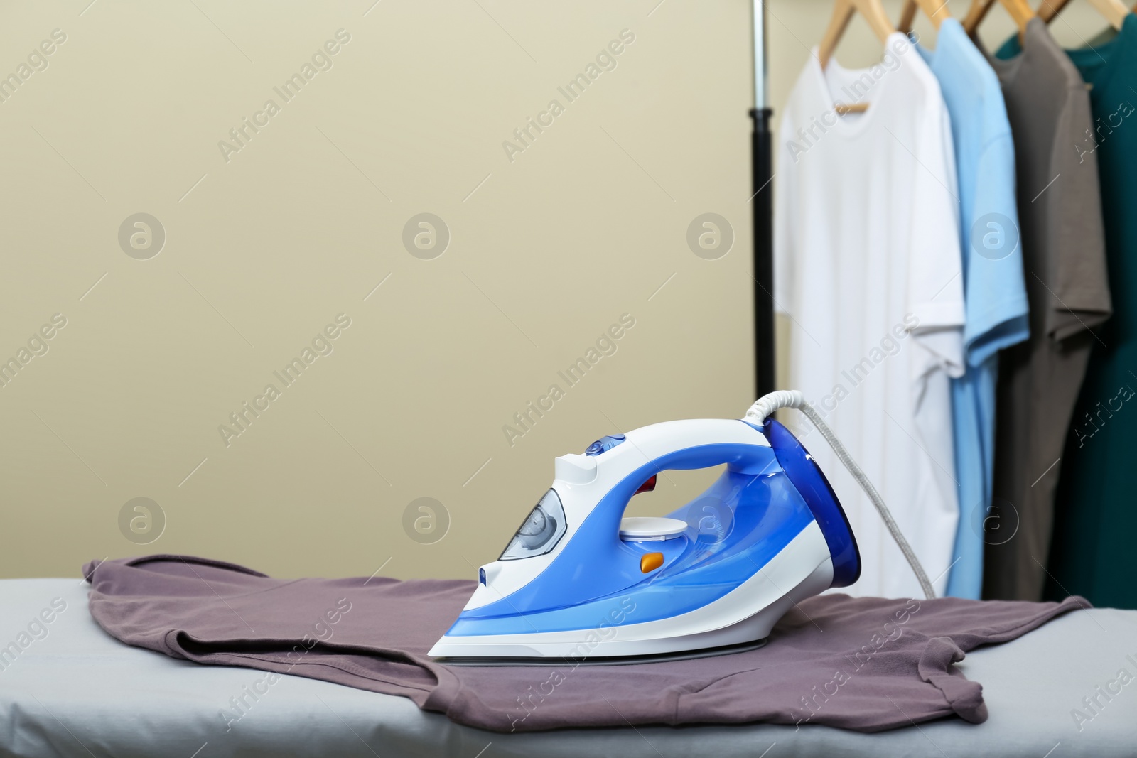 Photo of Modern iron on board and rack with clean clothes against beige background. Space for text