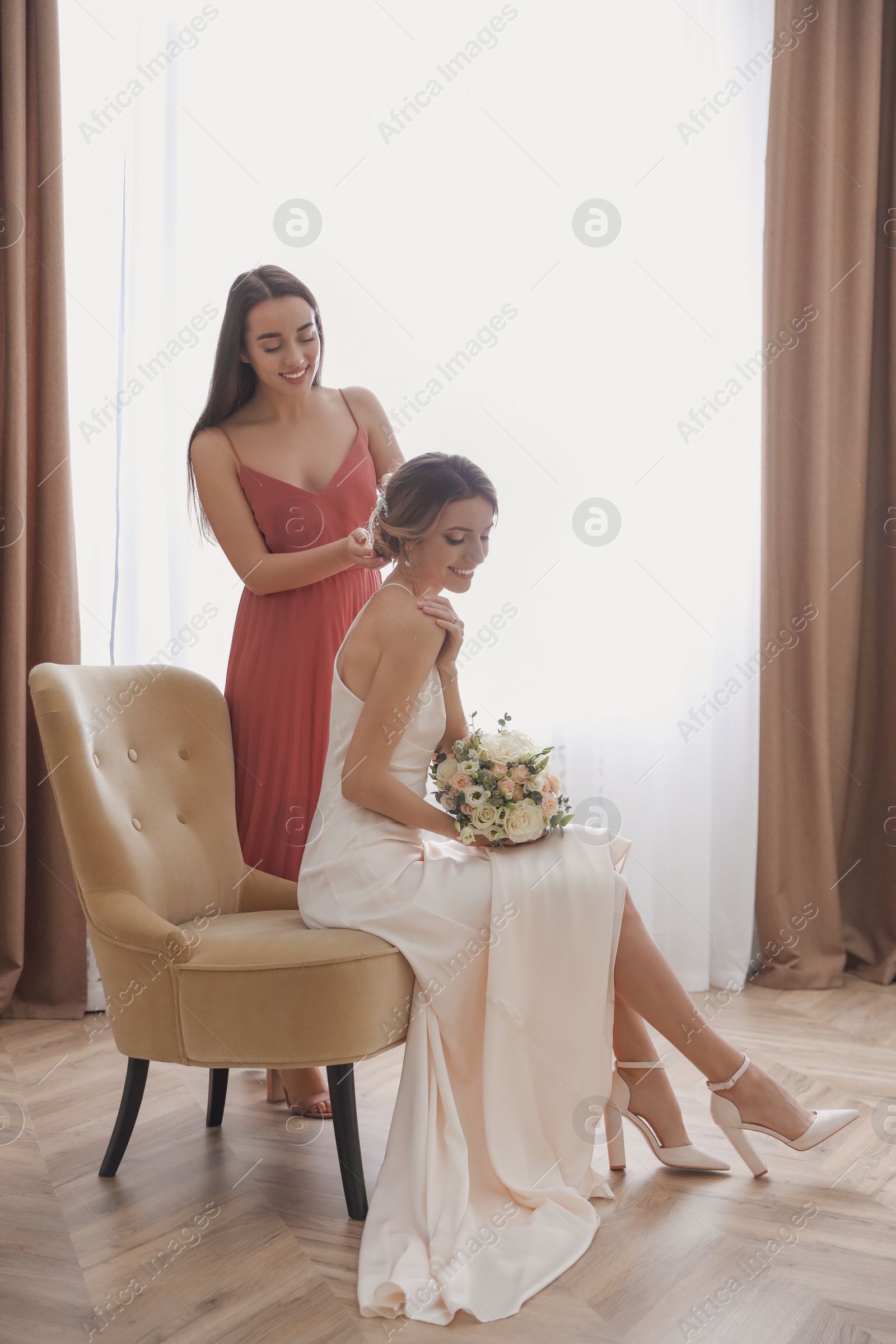 Photo of Gorgeous bride in beautiful wedding dress and her friend making hairstyle in room