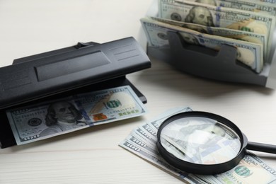 Photo of Modern currency detector with dollar banknotes and magnifying glass on white wooden table. Money examination device