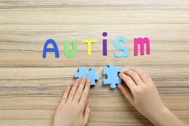 Woman putting together jigsaw puzzle pieces below word Autism on wooden table, top view