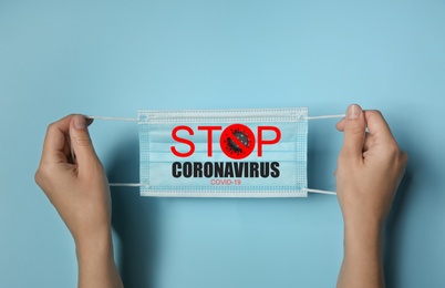 Image of Woman holding medical mask with text Stop Coronavirus on light blue background, closeup. Protective measures during pandemic