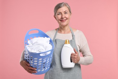 Happy housewife with detergent and basket full of laundry on pink background