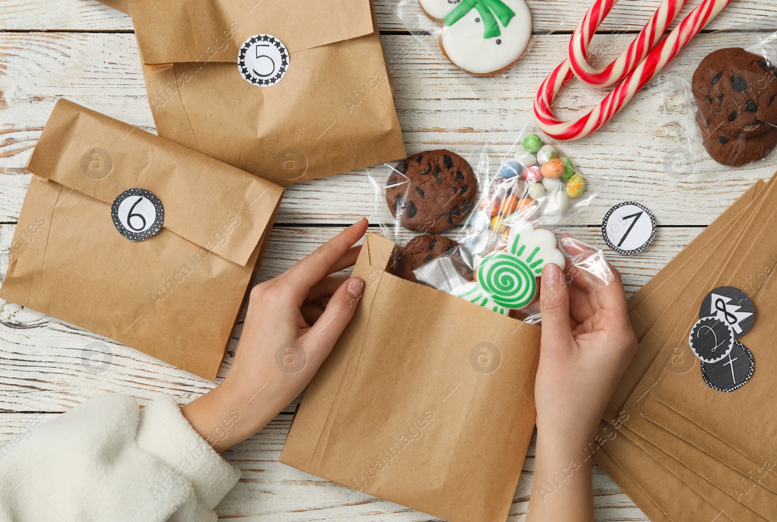 Photo of Woman putting Christmas treats into gift bag at white wooden table, top view. Creating advent calendar