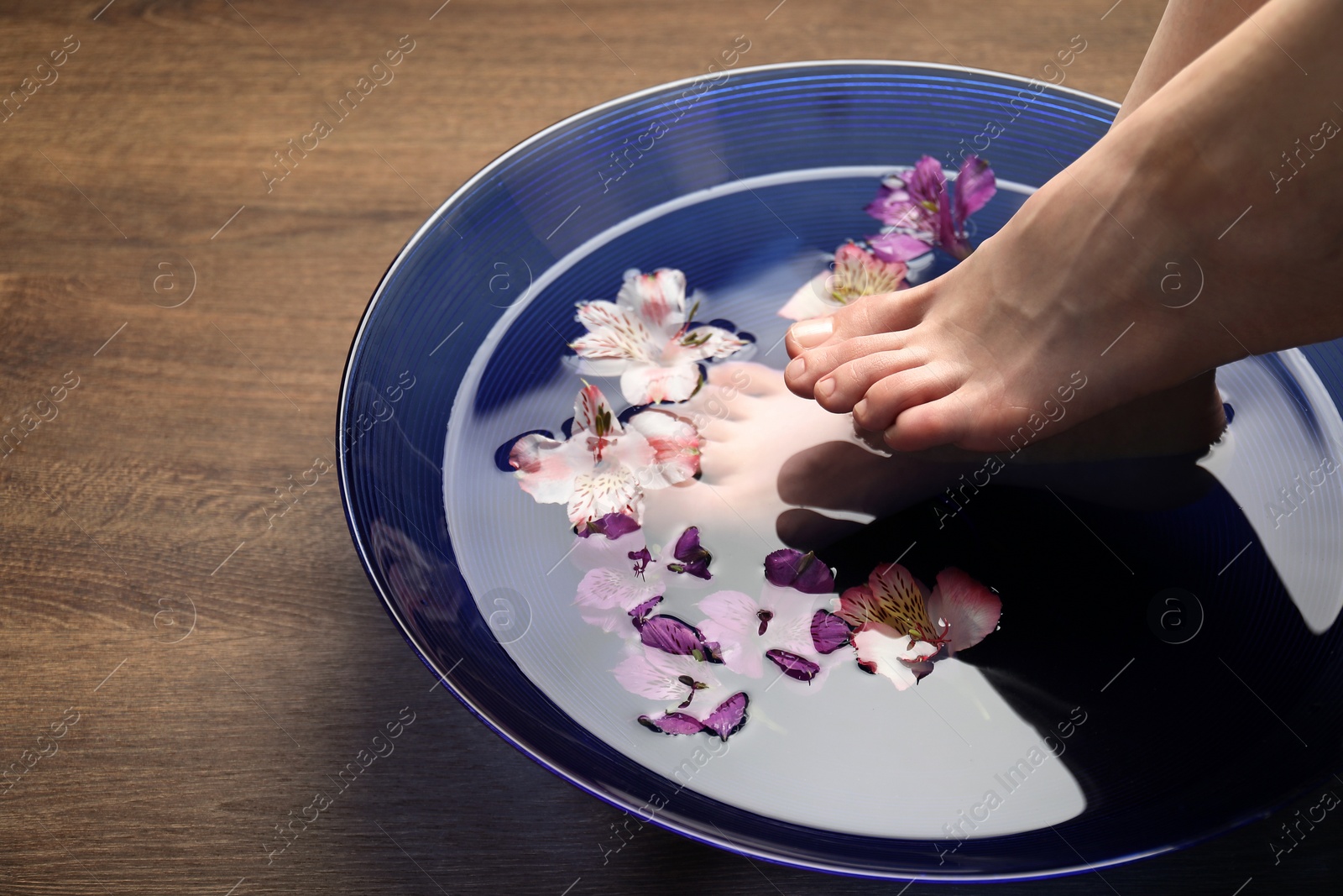 Photo of Woman soaking her feet in bowl with water and flowers on wooden floor, closeup with space for text. Spa treatment