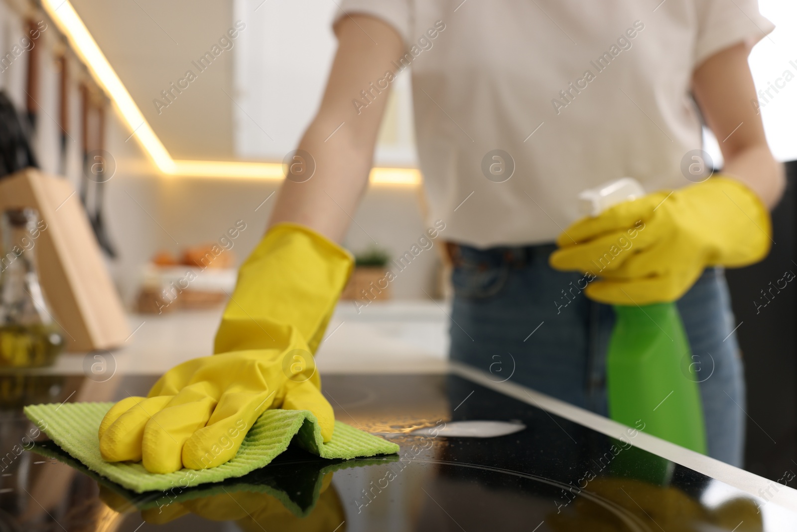 Photo of Woman with spray bottle and microfiber cloth cleaning electric stove in kitchen, closeup