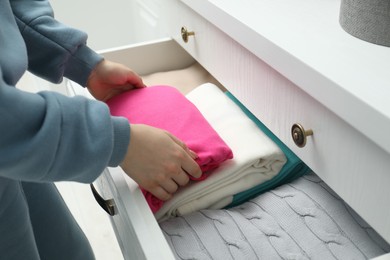 Photo of Woman putting folded clothes into drawer indoors, closeup