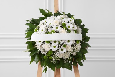 Photo of Funeral wreath of flowers with ribbon on wooden stand near white wall indoors