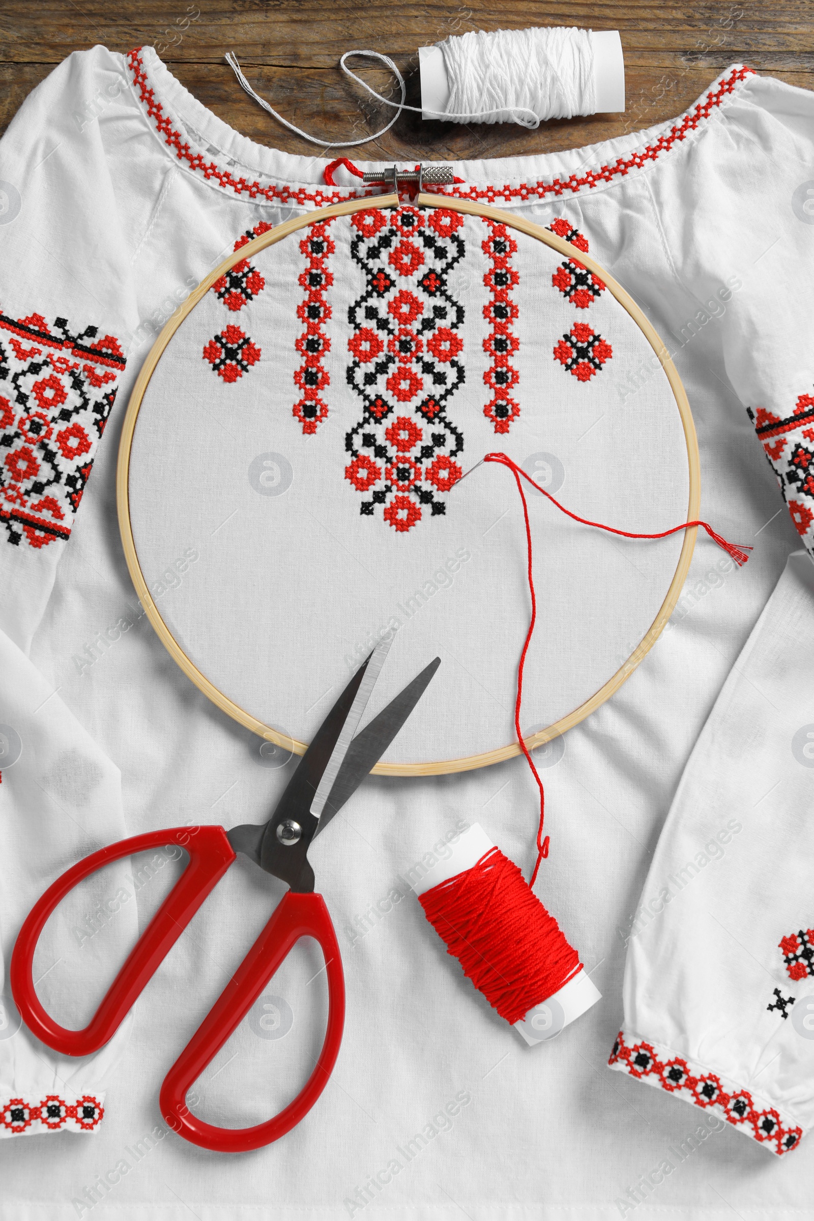 Photo of Shirt with red embroidery design in hoop, needle, scissors and threads on wooden table, flat lay. National Ukrainian clothes