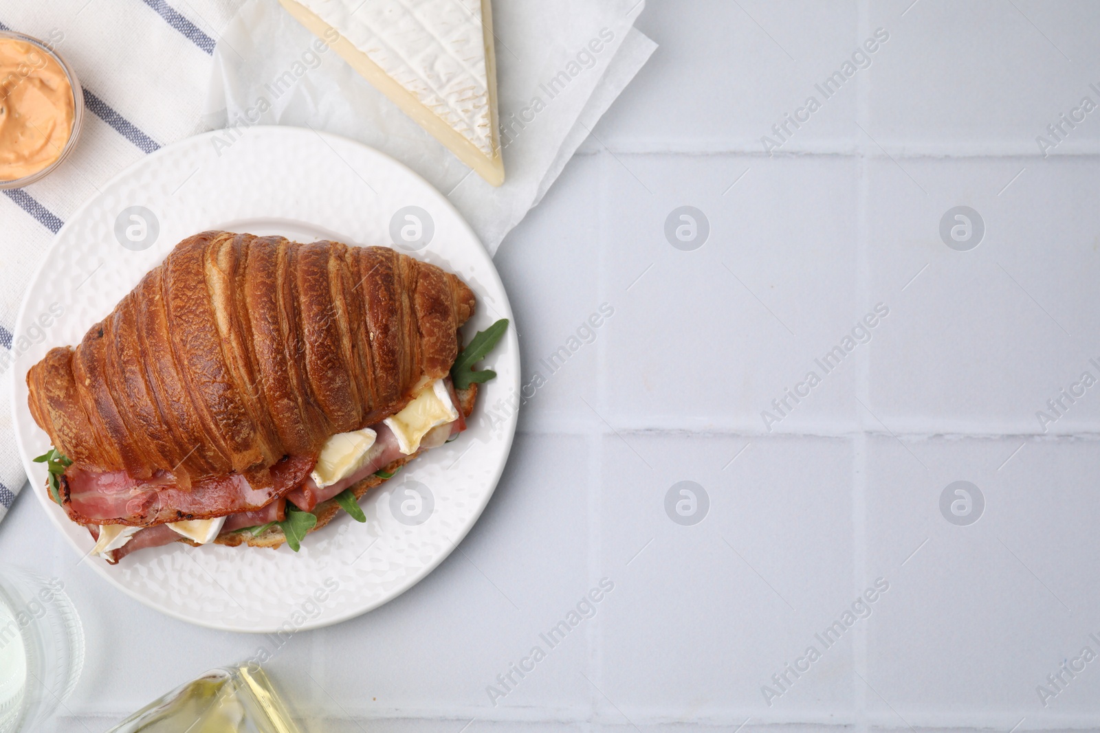 Photo of Tasty croissant with brie cheese, ham and bacon on white tiled table, flat lay. Space for text