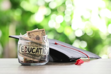 Photo of Glass jar with money and graduation hat on table against blurred background. Space for text
