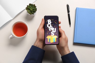 Image of Bonus gaining. Man holding smartphone at white table, top view. Illustration of open gift box, word and confetti on device screen
