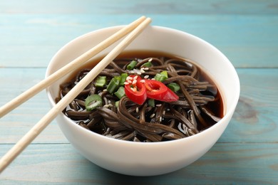 Photo of Tasty soup with buckwheat noodles (soba), chili pepper, green onion in bowl and chopsticks on light blue wooden table, closeup