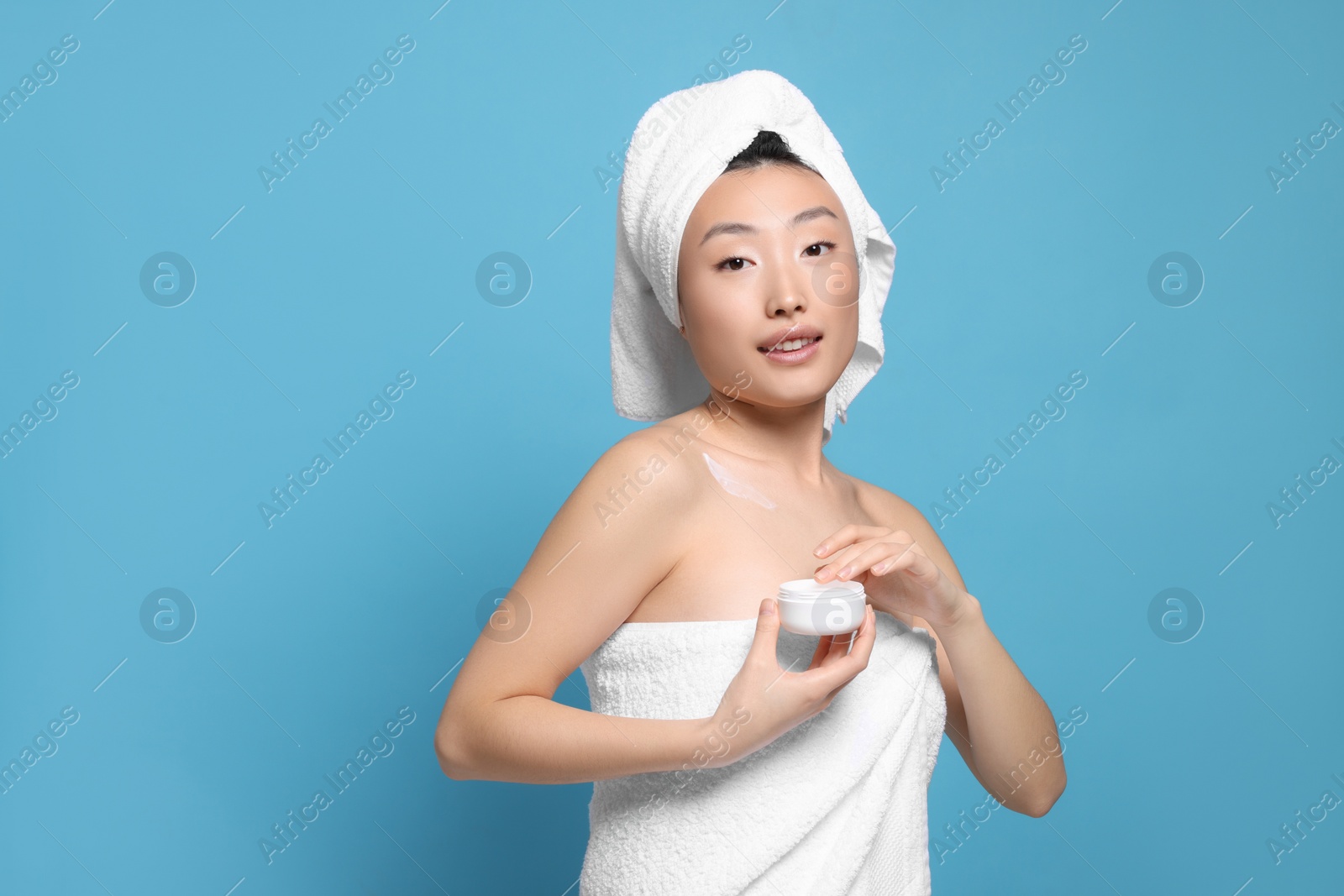 Photo of Beautiful young Asian woman with jar of body cream on light blue background, space for text