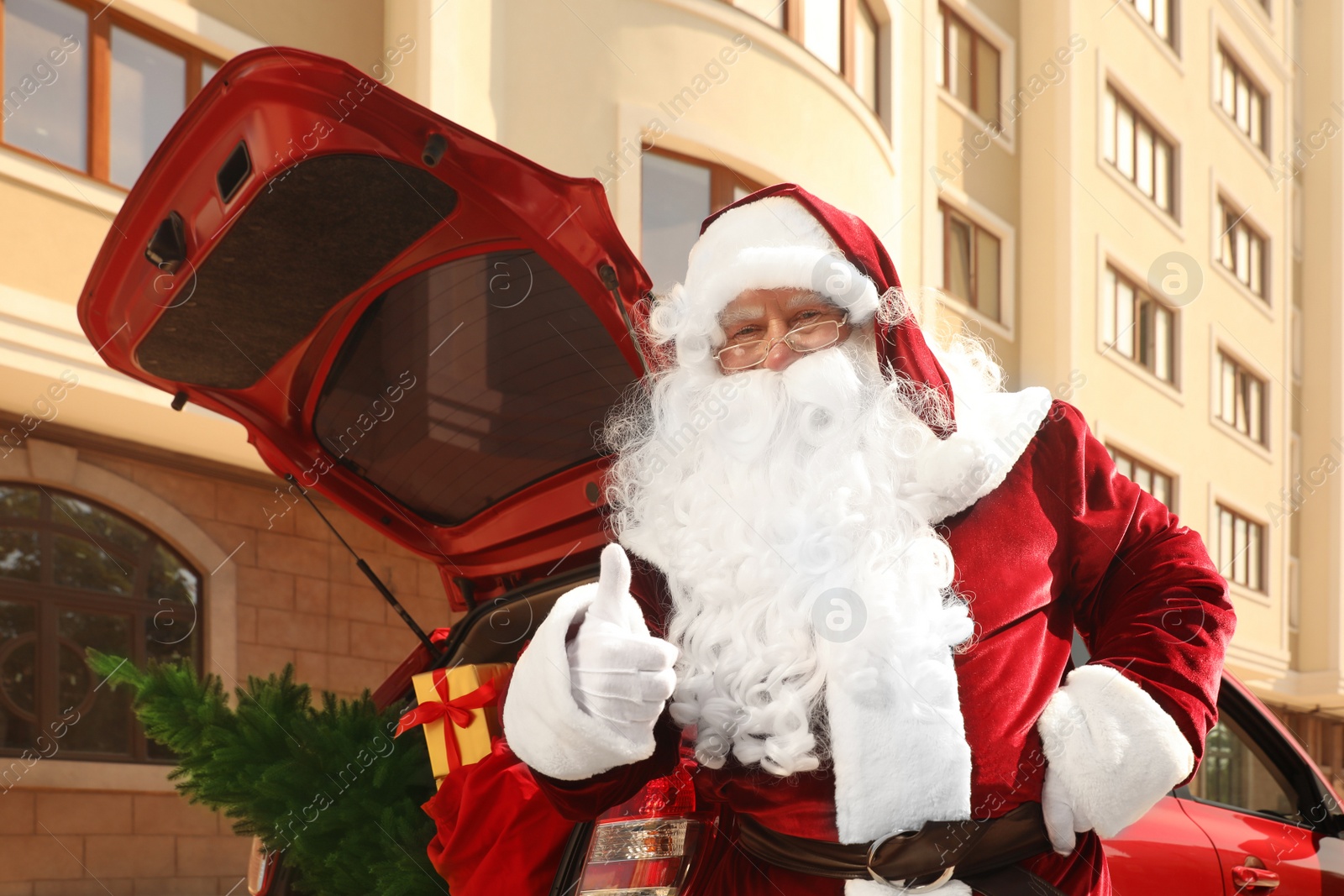 Photo of Authentic Santa Claus near car with fir tree and bag full of Christmas gifts in open trunk outdoors