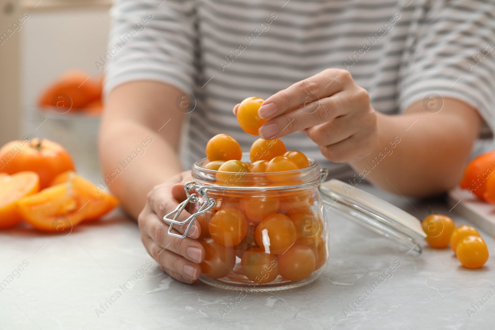 Photo of Woman putting yellow tomatoes into glass jar at light kitchen table, closeup. Pickling vegetables