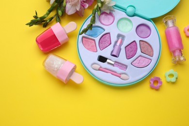 Decorative cosmetics for kids. Eye shadow palette, lipsticks, accessories and flowers on yellow background, flat lay. Space for text