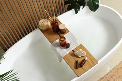 Wooden bath tray with open book, candle and body care products on tub indoors. Relaxing atmosphere