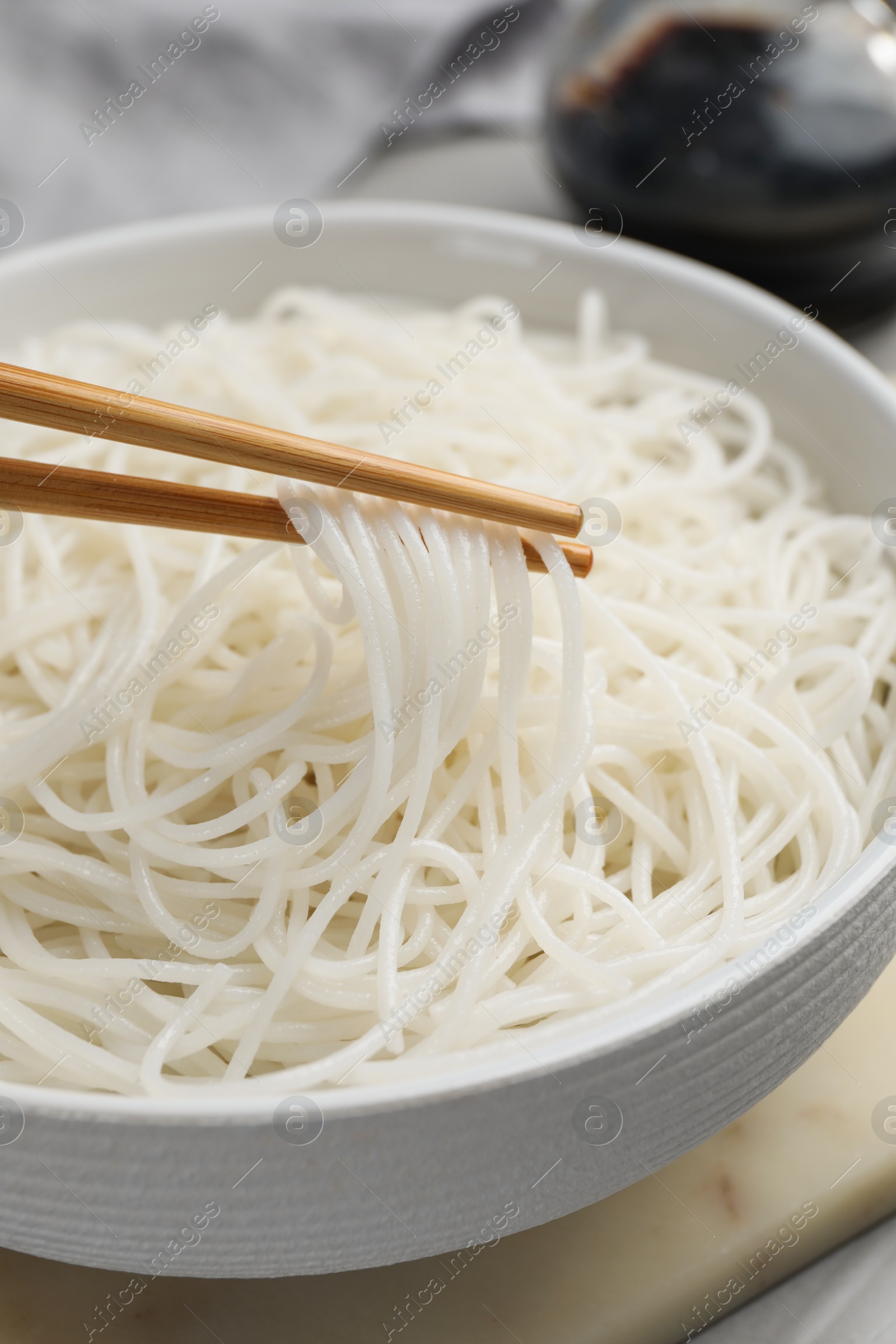 Photo of Chopsticks with cooked rice noodles over bowl on table, closeup