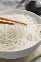 Chopsticks with cooked rice noodles over bowl on table, closeup