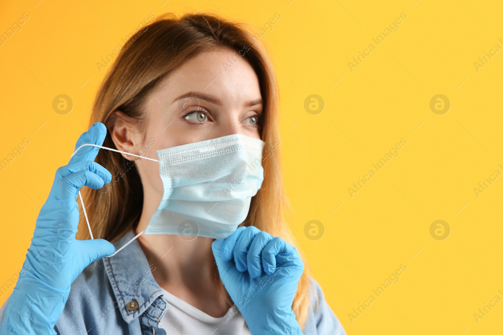 Photo of Young woman in medical gloves putting on protective face mask against yellow background. Space for text