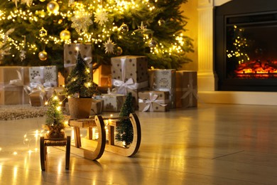 Wooden decorative sleighs in front of many gift boxes, festive lights and Christmas tree on floor. Space for text