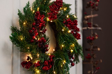 Beautiful Christmas wreath with red berries and fairy lights hanging on white door, closeup. Space for text