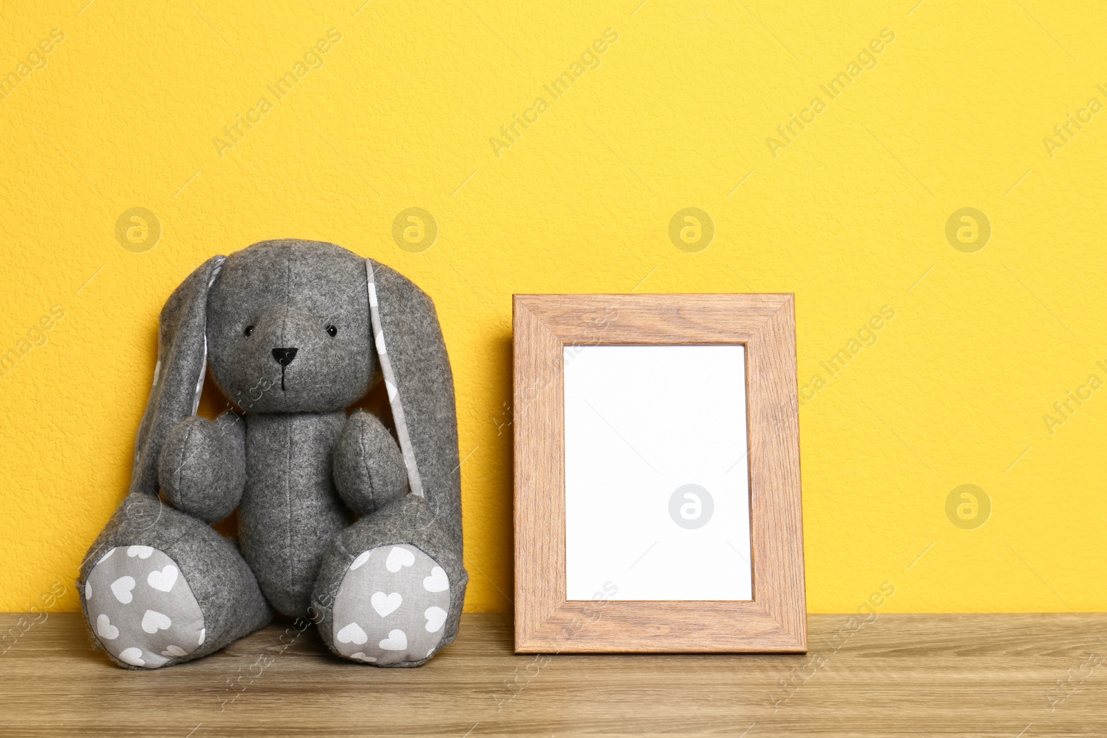 Photo of Empty photo frame and and soft rabbit on wooden table against yellow background, space for text. Child room interior