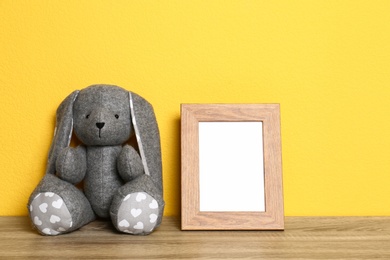 Photo of Empty photo frame and and soft rabbit on wooden table against yellow background, space for text. Child room interior