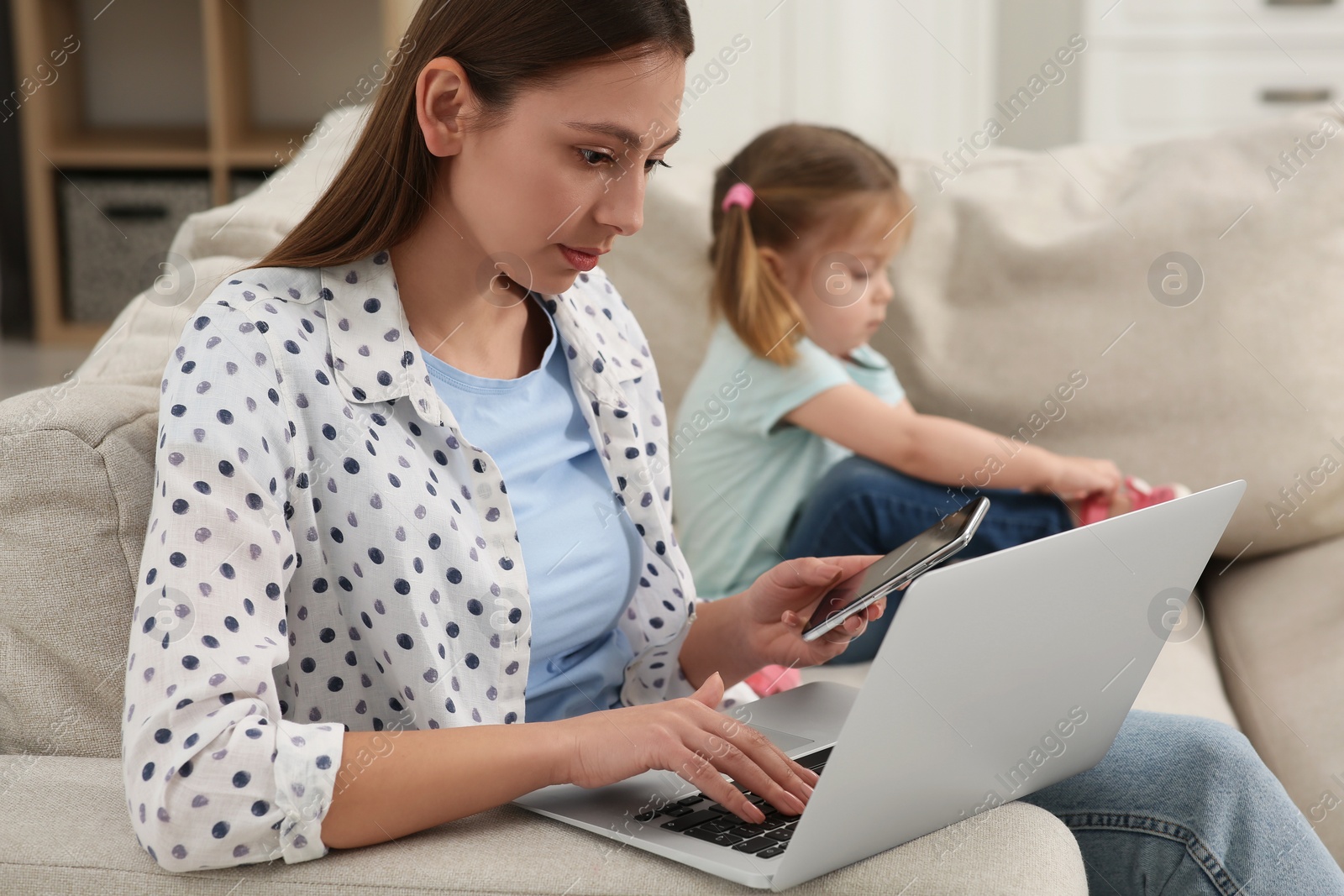 Photo of Woman with laptop working remotely at home. Mother and daughter on sofa in living room