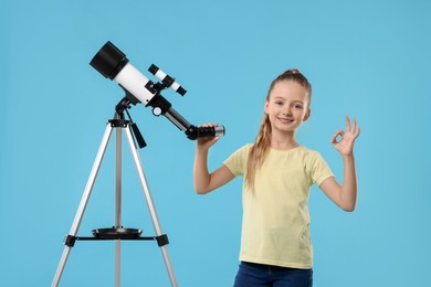 Photo of Happy little girl with telescope showing ok gesture on light blue background