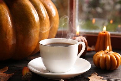 Photo of Cup of hot drink and pumpkin shaped candles on wooden table near window, closeup. Cozy autumn atmosphere