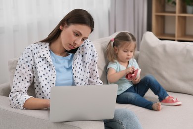 Photo of Working remotely at home. Busy mother with laptop talking on phone. Woman and her daughter on sofa in living room