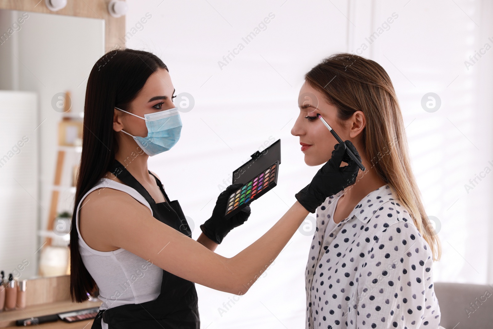 Photo of Makeup artist in protective mask and gloves working with woman indoors. Preventive measures during COVID-19 pandemic