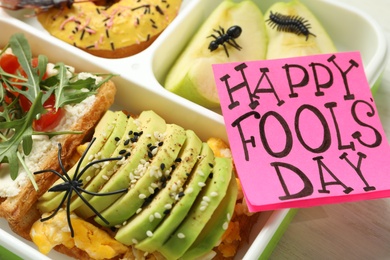 Photo of Lunchbox with fake spider, bugs and Happy Fools' Day note on table, closeup