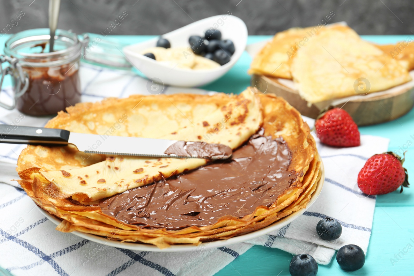 Photo of Tasty crepes with chocolate paste and berries served on turquoise wooden table, closeup