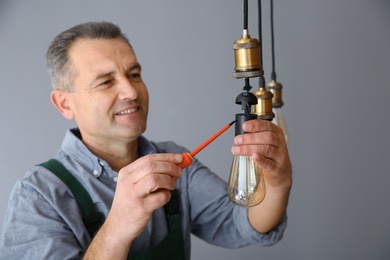 Electrician with screwdriver repairing ceiling lamp on grey background