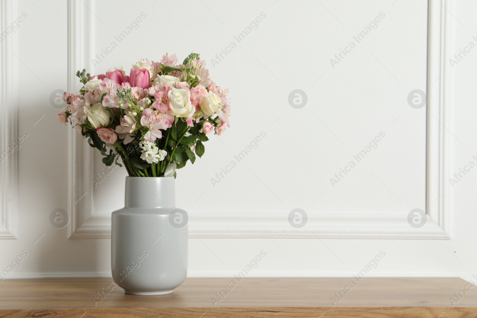 Photo of Beautiful bouquet of fresh flowers in vase on wooden table near white wall, space for text