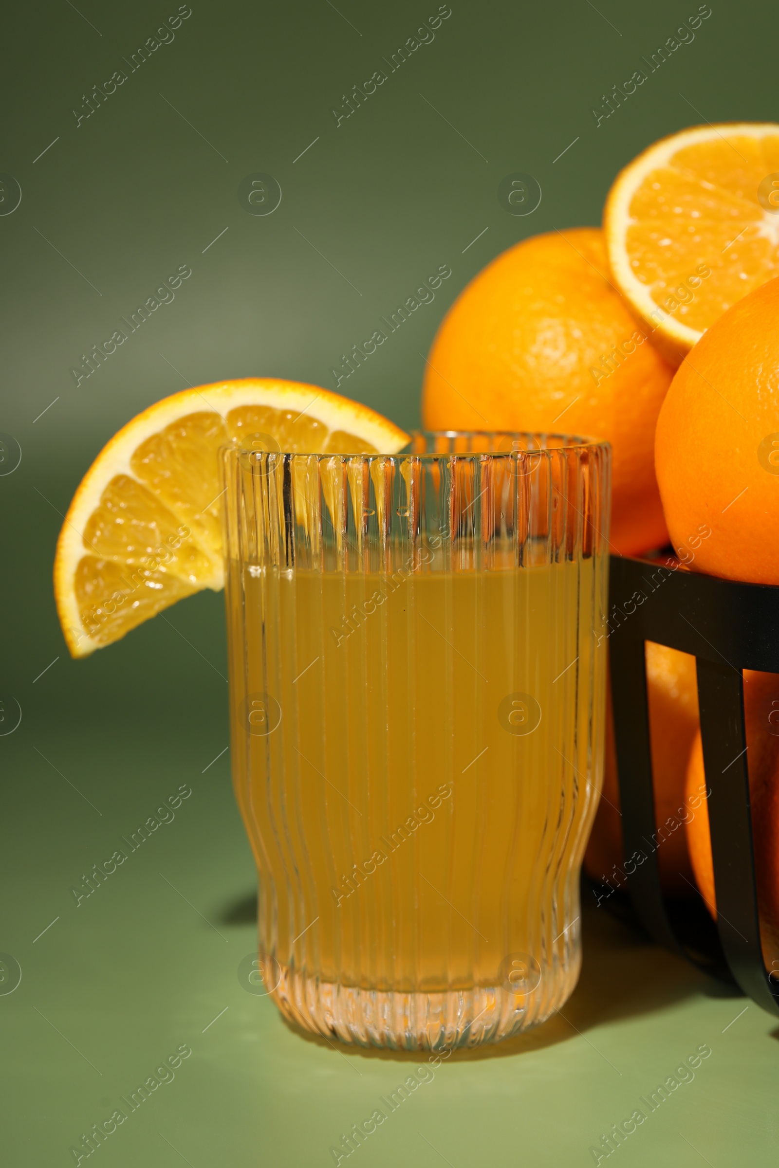 Photo of Fresh oranges in metal basket and glass of juice on green background, closeup