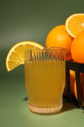 Fresh oranges in metal basket and glass of juice on green background, closeup