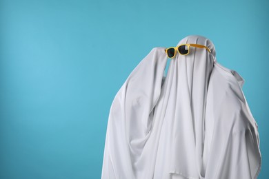 Photo of Stylish ghost. Person covered with white sheet in sunglasses on light blue background, space for text